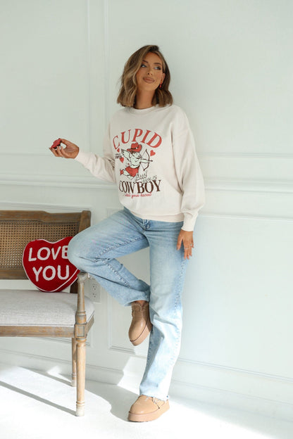 A woman in a Light Beige Cupid Cowboy Graphic Sweatshirt and jeans stands by a bench with a red LOVE YOU pillow, holding a small object.