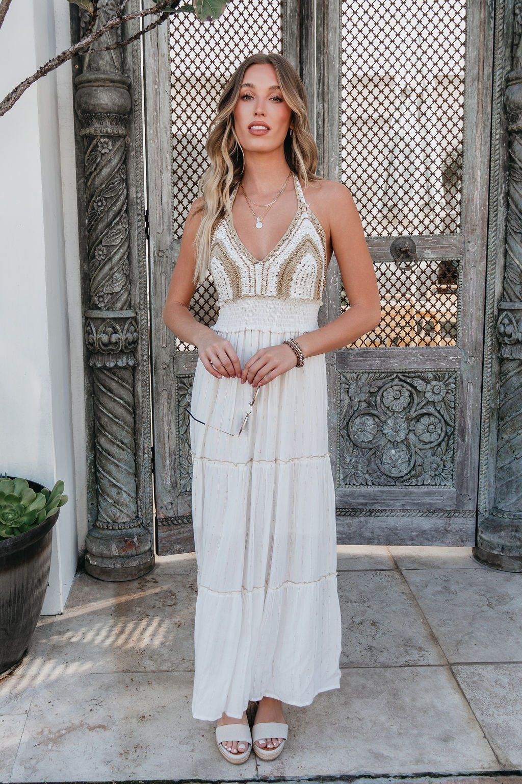 Woman in the Ivory Drift Smock Tiered Maxi Dress stands before ornate wooden doors, holding sunglasses.