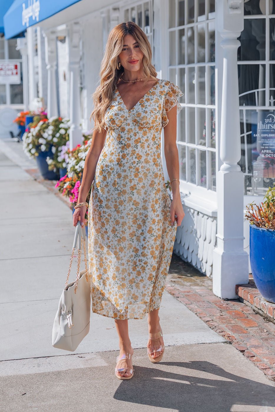 A woman in an Ivory Floral Chiffon Midi Dress walks along a sidewalk with a cream handbag, flowers and shops behind her.