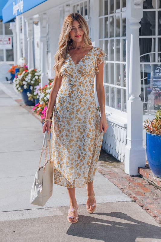 A woman in an Ivory Floral Chiffon Midi Dress walks along a sidewalk with a cream handbag, flowers and shops behind her.