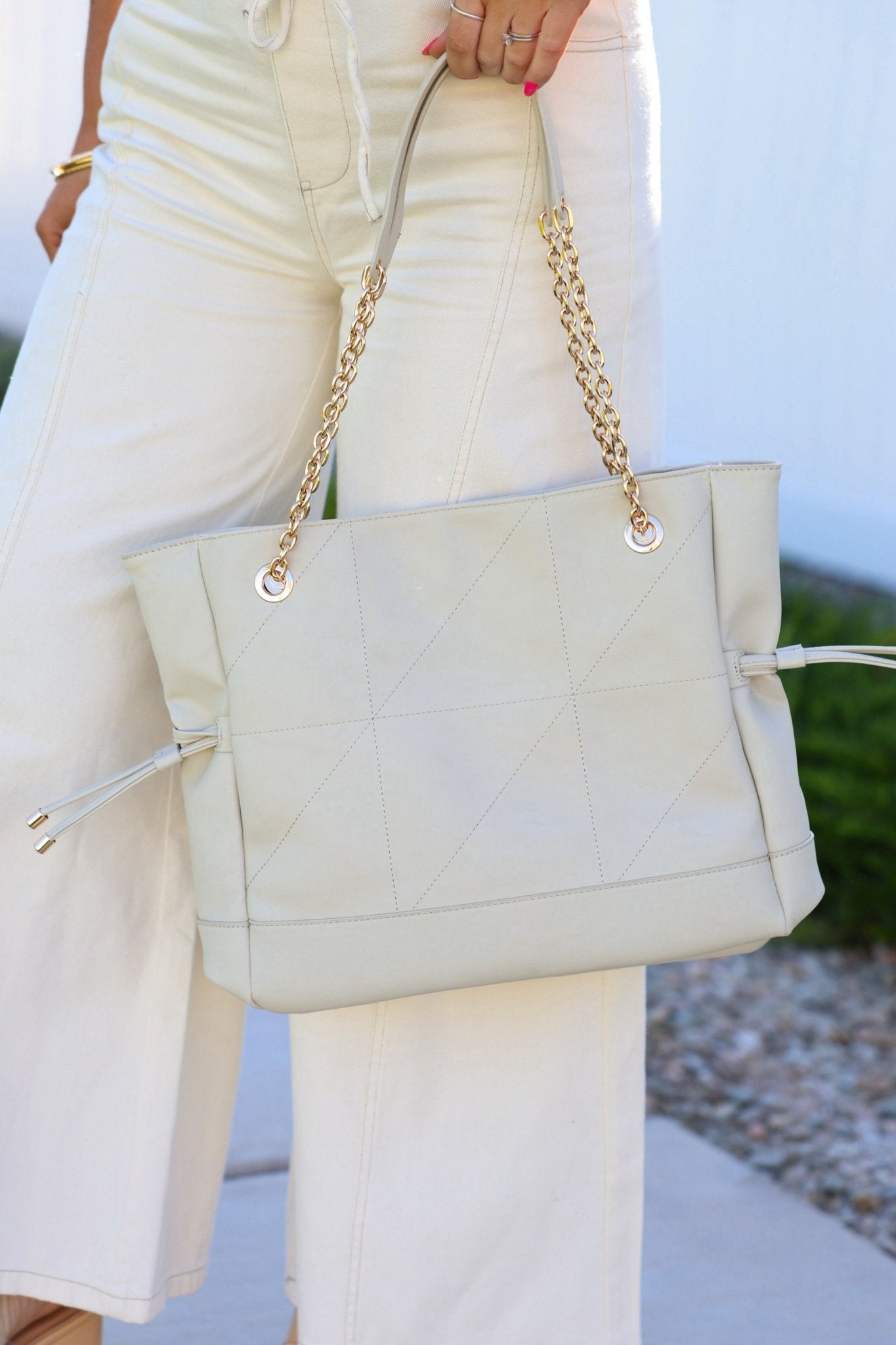 A person in off-white pants holds the Ivory Kinsley Embellished Tote Bag with gold chain straps, ideal for everyday use.