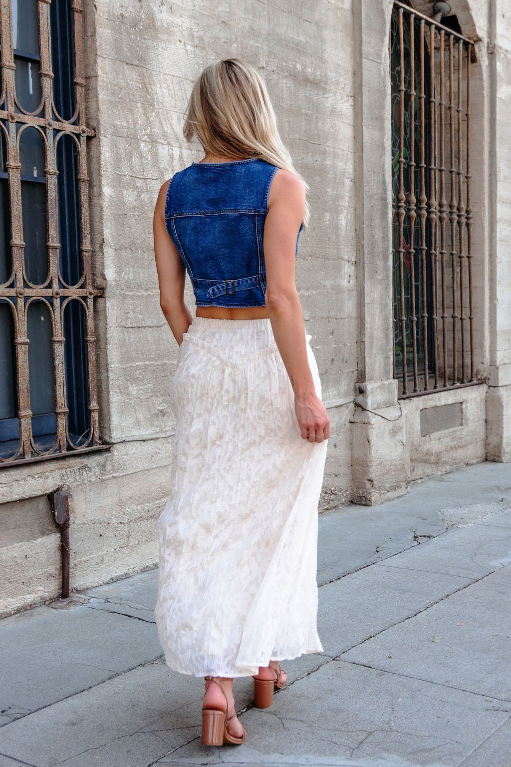 A woman with blonde hair in a denim vest and Ivory Lace Ruffled Maxi Skirt walks on a city sidewalk by barred windows.