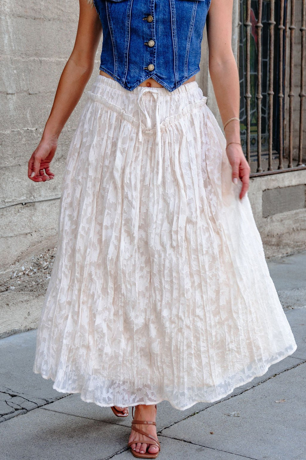 Woman in a sleeveless blue denim vest and Ivory Lace Ruffled Maxi Skirt, walking beside a concrete wall.