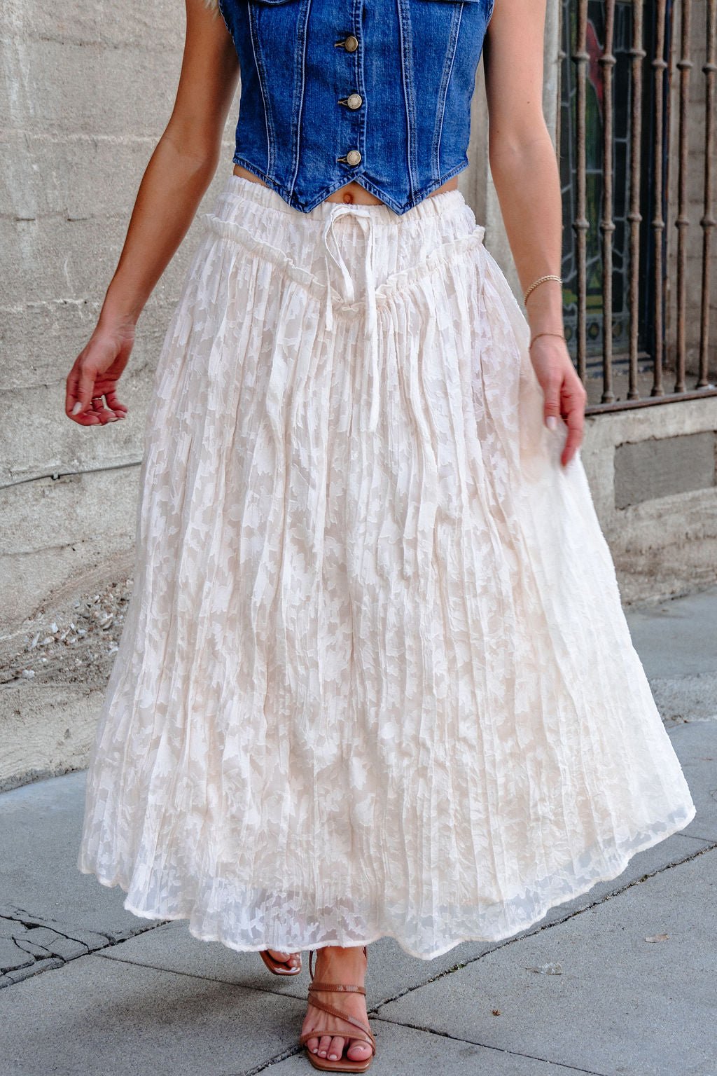Woman in a sleeveless blue denim vest and Ivory Lace Ruffled Maxi Skirt, walking beside a concrete wall.