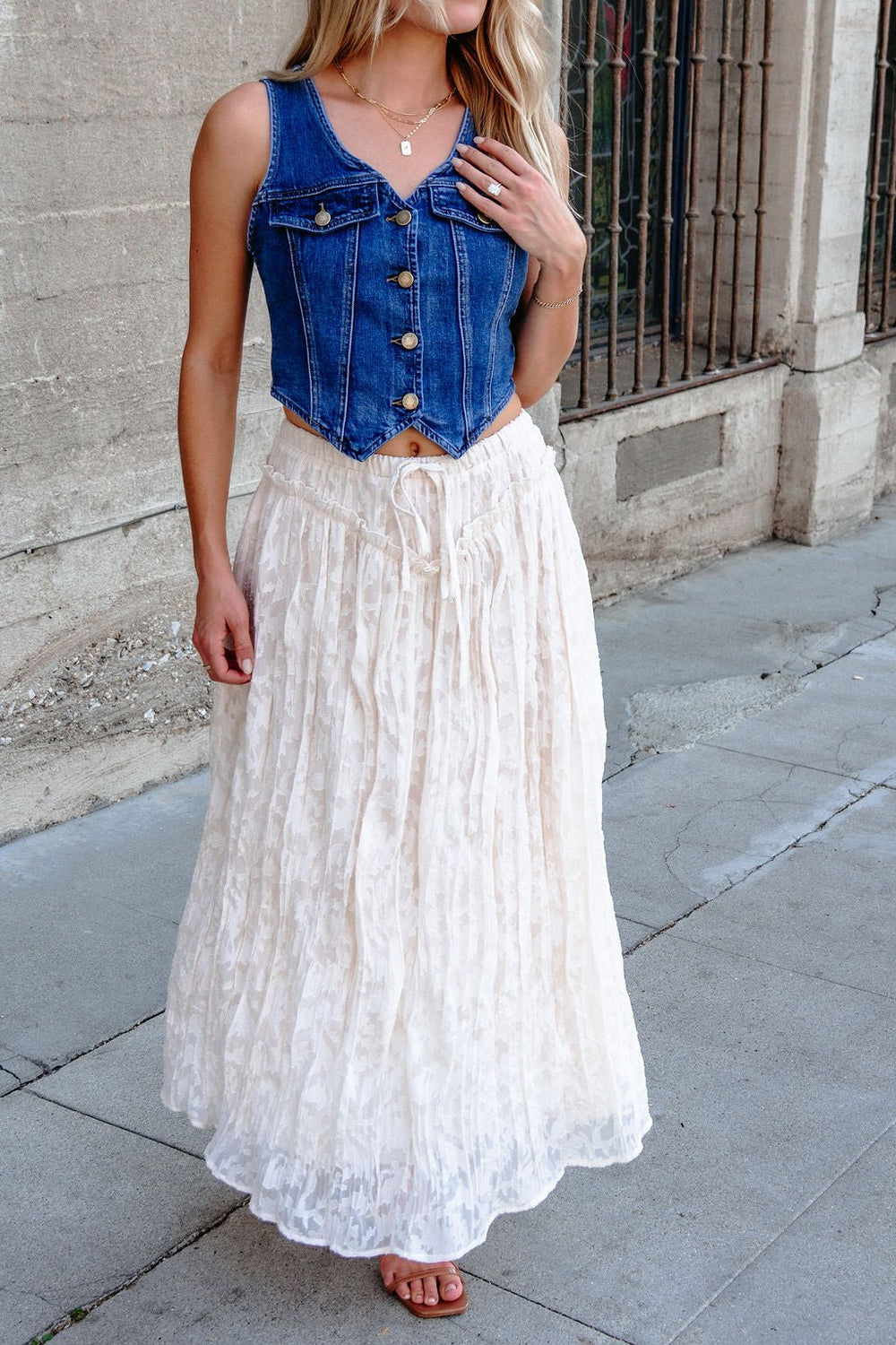 Woman wears an Ivory Lace Ruffled Maxi Skirt - FINAL SALE, standing on a city sidewalk near a concrete wall and iron fence.