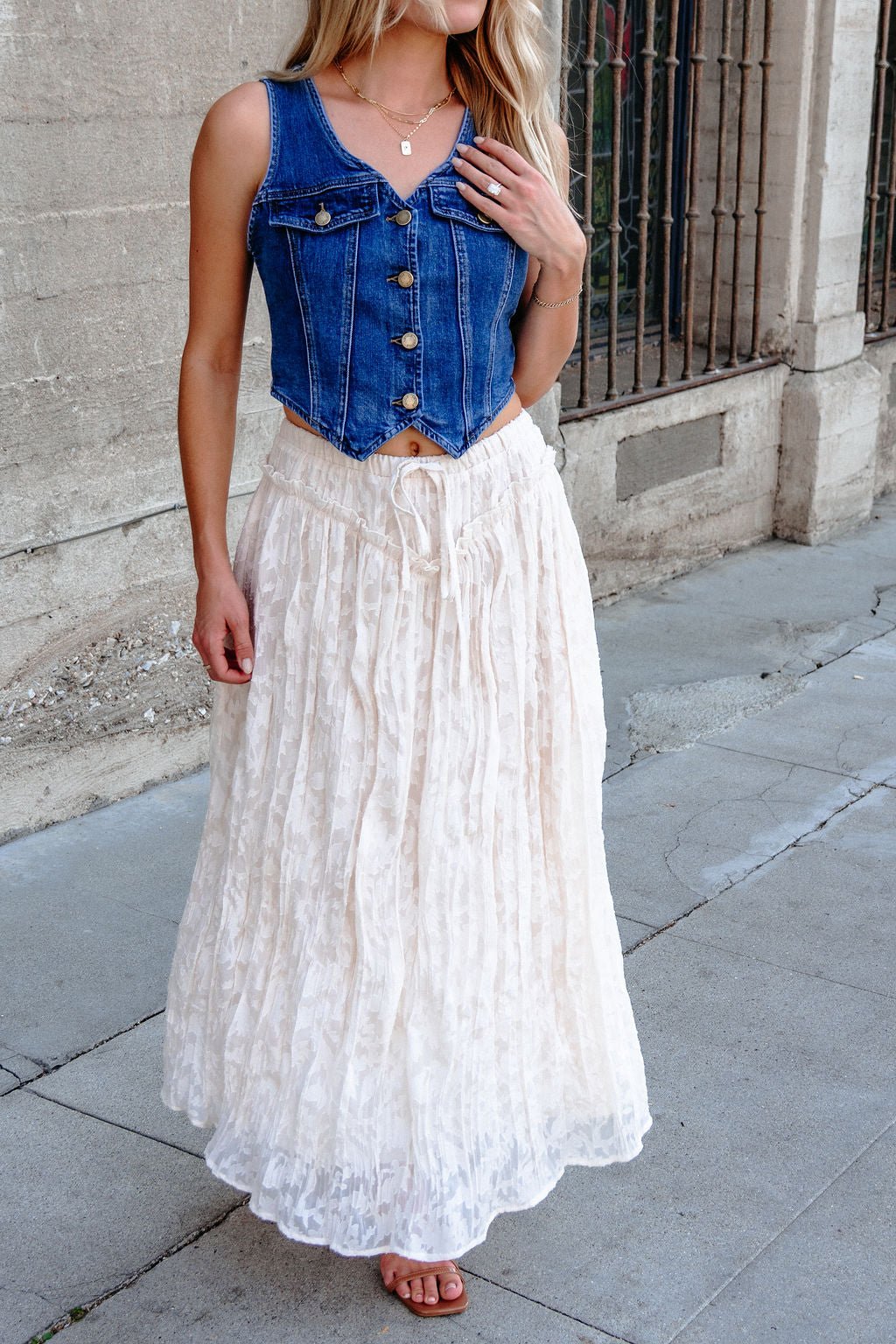 Woman wears an Ivory Lace Ruffled Maxi Skirt - FINAL SALE, standing on a city sidewalk near a concrete wall and iron fence.
