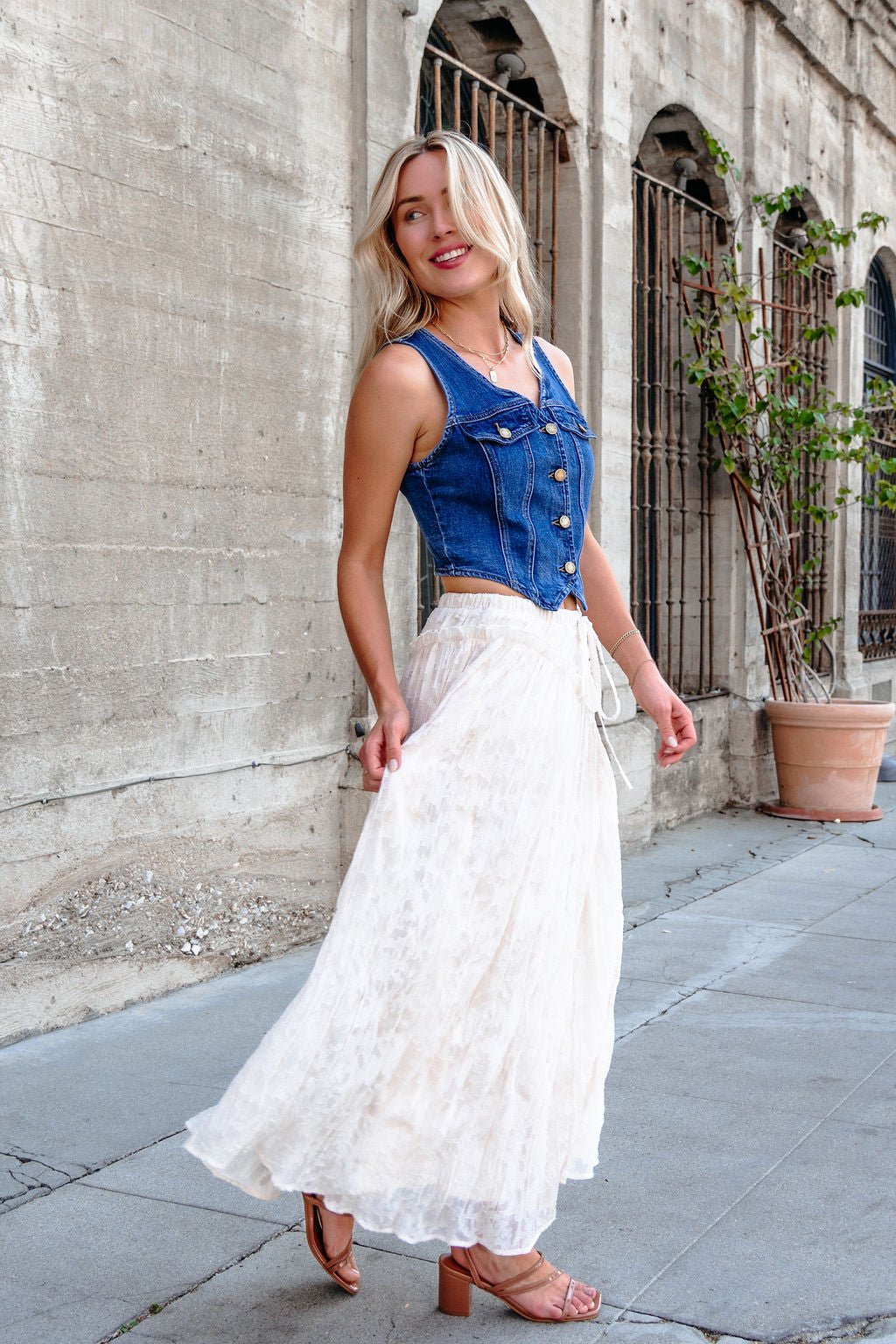 Woman in a denim sleeveless top and Ivory Lace Ruffled Maxi Skirt - FINAL SALE smiles while walking by a stone building.