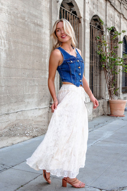 Woman in a denim sleeveless top and Ivory Lace Ruffled Maxi Skirt - FINAL SALE smiles while walking by a stone building.