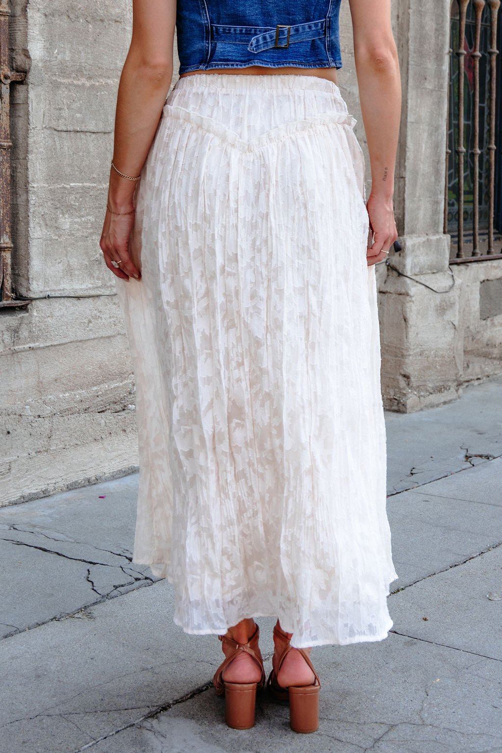 A person in a blue denim vest, pink platform sandals, and an Ivory Lace Ruffled Maxi Skirt stands on a stone sidewalk.