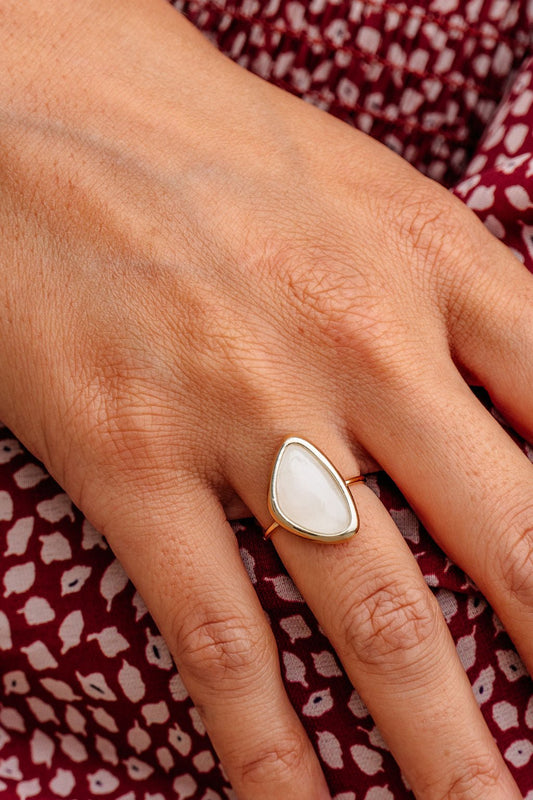 A hand wears the Ivory Orante Resin Stone Acrylic Ring, featuring a large white stone, resting on maroon patterned fabric.