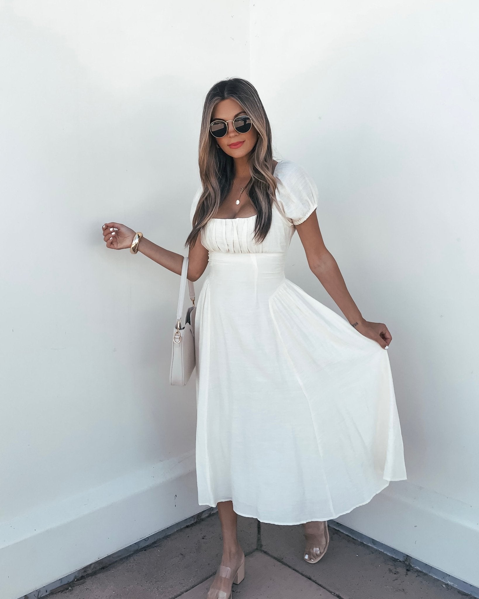 Woman wearing the Ivory Puff Sleeve Shirred Midi Dress and sunglasses poses against a white wall, holding her dress.