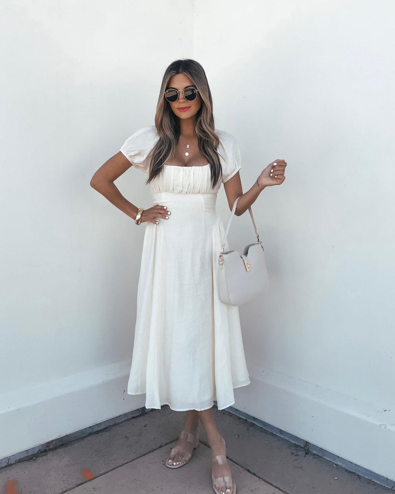 Woman in an Ivory Puff Sleeve Shirred Midi Dress, sunglasses, and sandals holds a beige handbag and smiles against a white wall.
