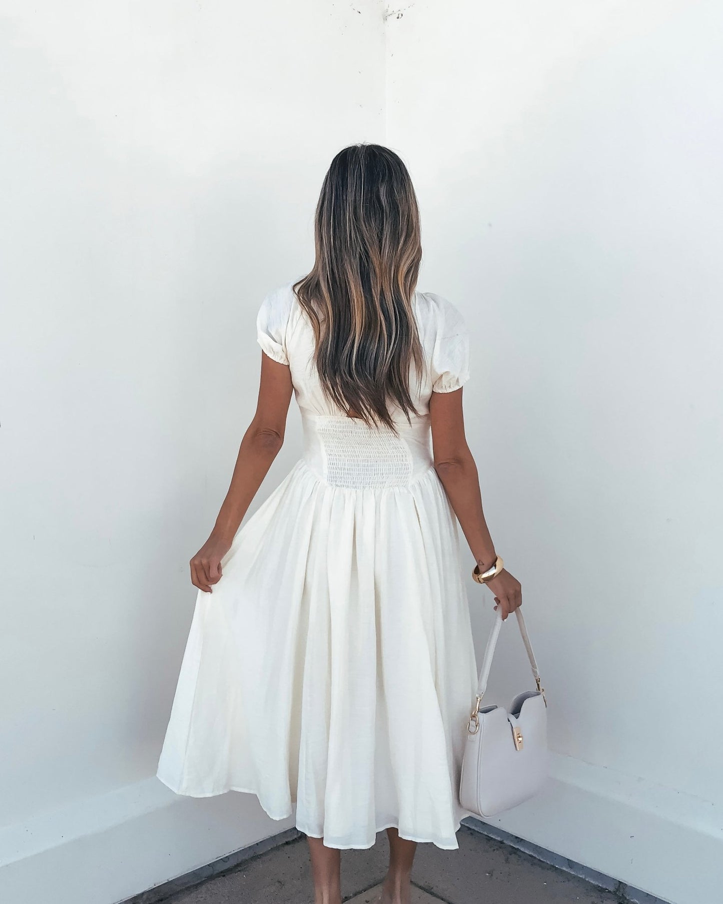 A woman in an Ivory Puff Sleeve Shirred Midi Dress faces a white wall, holding a light purse at her side.