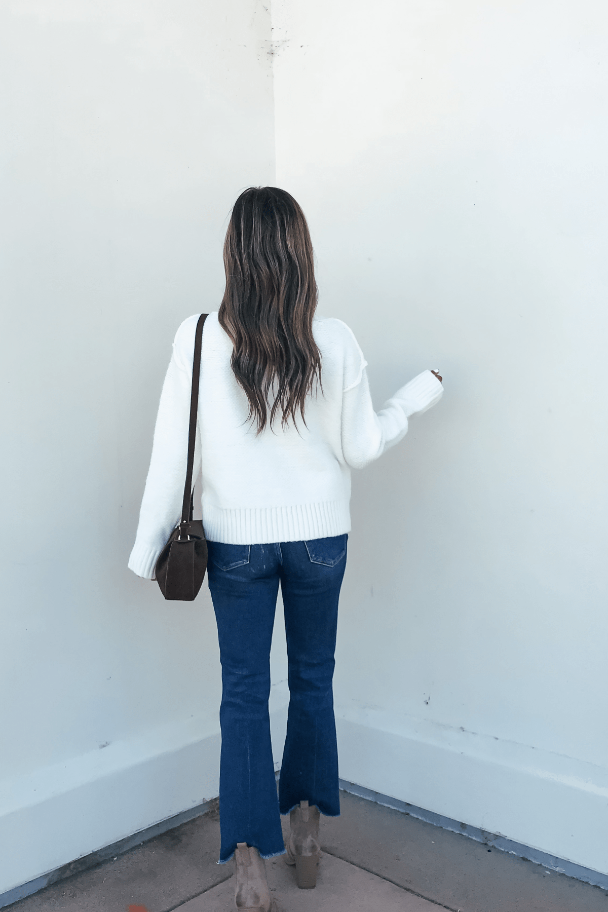 A woman in an Ivory Seam Detail Ribbed Sweater, blue jeans, heels, and a brown crossbody bag stands facing a white wall.