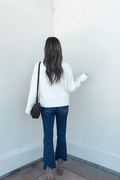 A woman in an Ivory Seam Detail Ribbed Sweater, blue jeans, heels, and a brown crossbody bag stands facing a white wall.