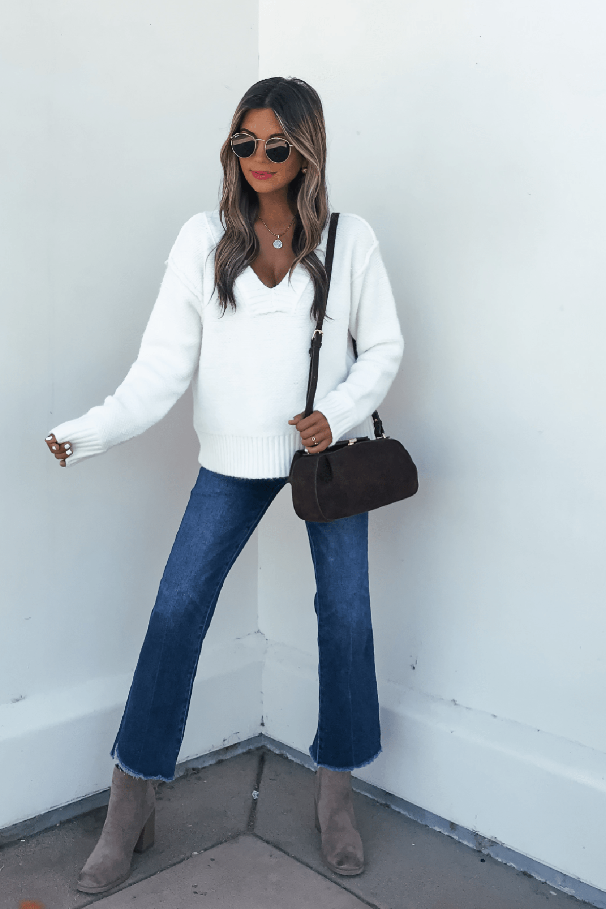 Woman in sunglasses wears an Ivory Seam Detail Ribbed Sweater, blue jeans, taupe boots, and carries a dark handbag against a white wall.