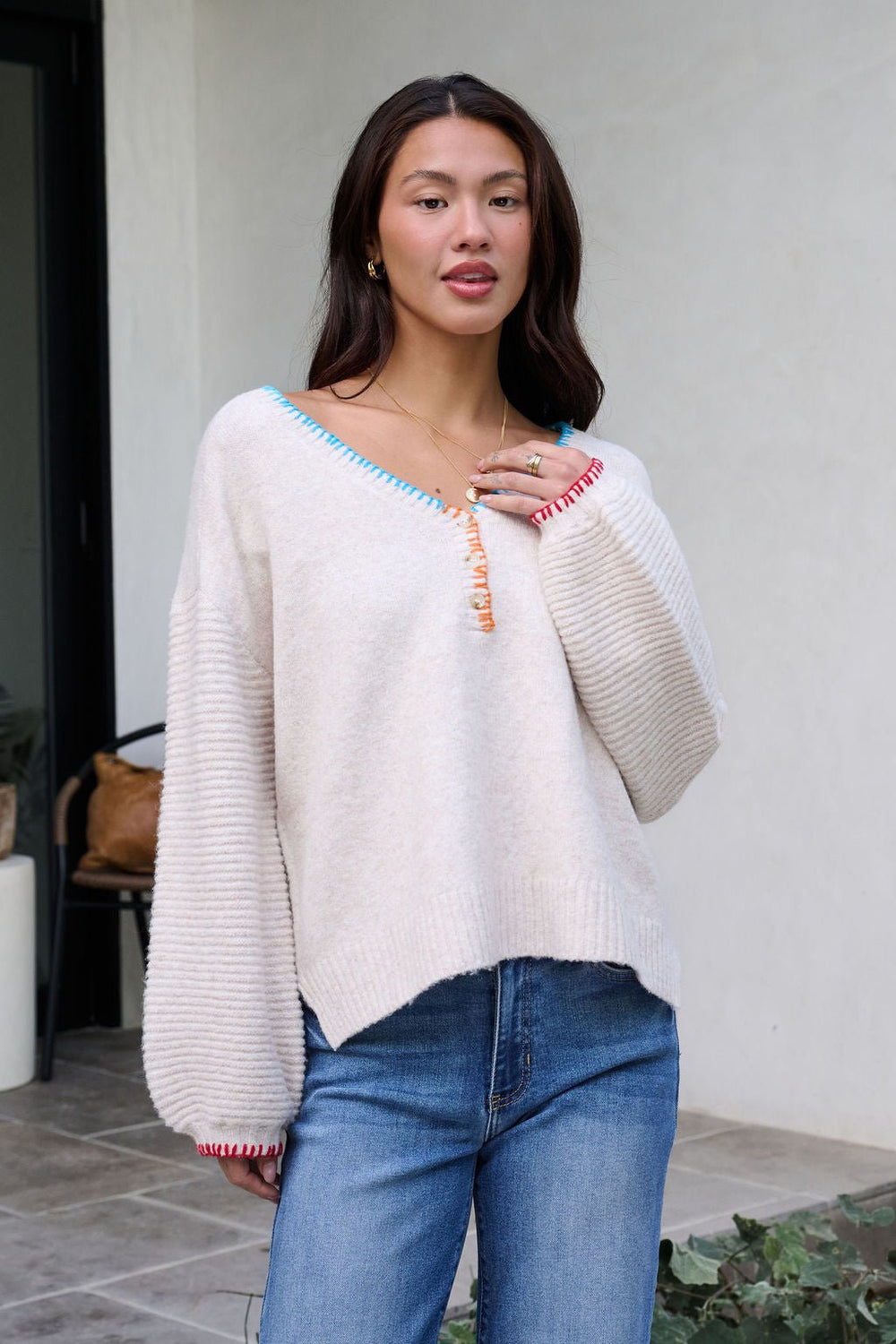 A woman in an Ivory Stitch Trim Button Up Sweater - FINAL SALE and blue jeans stands outdoors, holding the sweater near her collarbone.