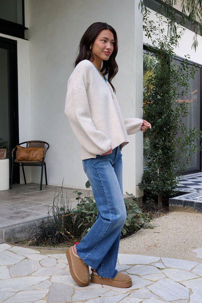 A woman in an Ivory Stitch Trim Button Up Sweater, blue jeans, and tan slippers smiles outdoors on a stone patio.