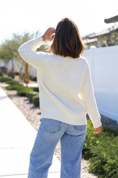 A woman in an Ivory Textured Cable Knit Sweater and blue jeans stands outdoors, facing away, touching her hair.