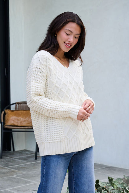 A woman wearing the Ivory Textured Cable Knit Sweater and blue jeans smiles outdoors near a chair and greenery.