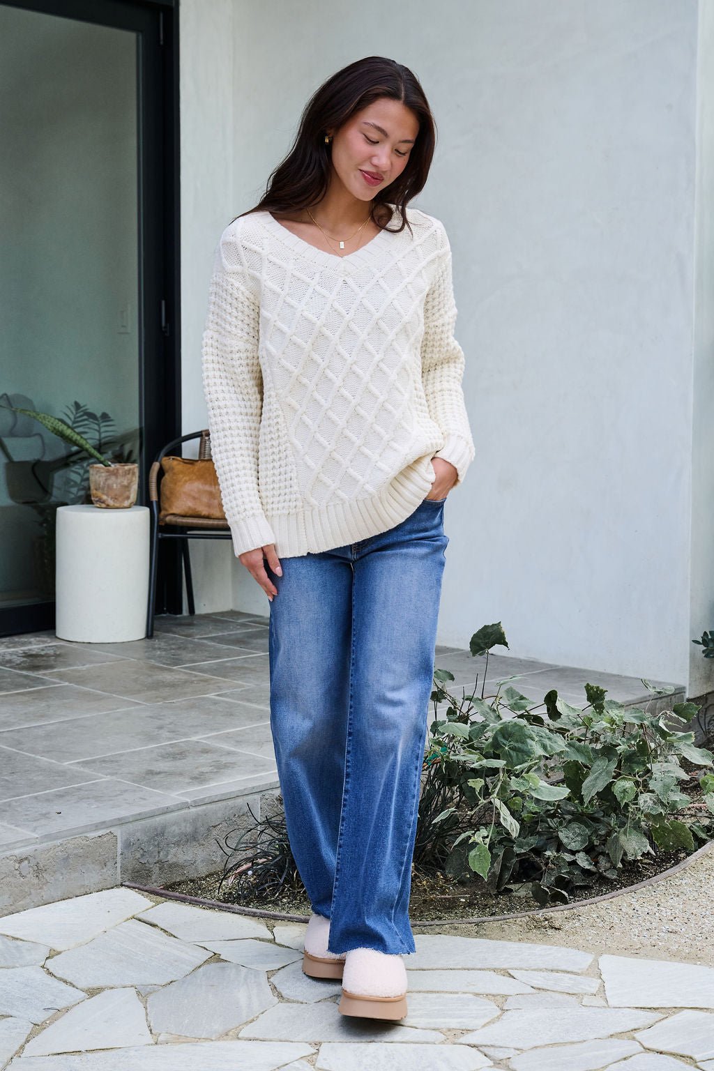 A woman in an Ivory Textured Cable Knit Sweater and blue jeans stands outside a modern home, smiling as she looks down.