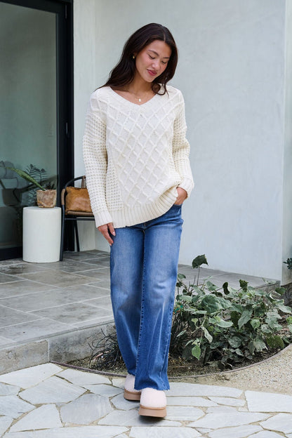 A woman in an Ivory Textured Cable Knit Sweater and blue jeans stands outside a modern home, smiling as she looks down.