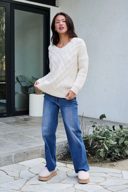 A woman wears the Ivory Textured Cable Knit Sweater, blue jeans, and slippers by a modern patio with plants and a glass door.