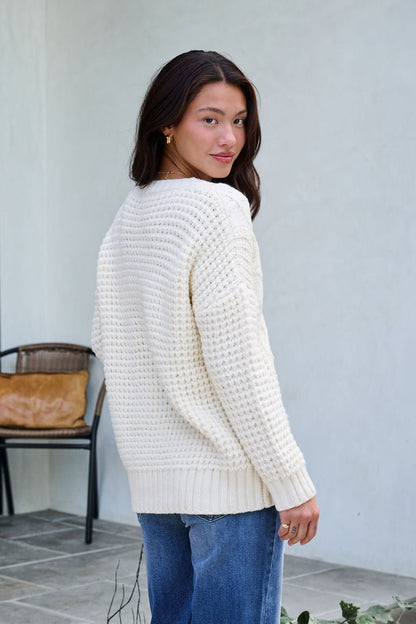 Woman with long brown hair wearing an Ivory Textured Cable Knit Sweater and blue jeans, standing outdoors looking over her shoulder.