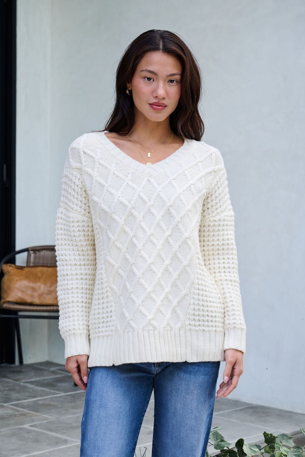 A woman wears the Ivory Textured Cable Knit Sweater and blue jeans outside on a tiled patio, plants and a chair behind her.