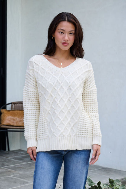 A woman wears the Ivory Textured Cable Knit Sweater and blue jeans outside on a tiled patio, plants and a chair behind her.
