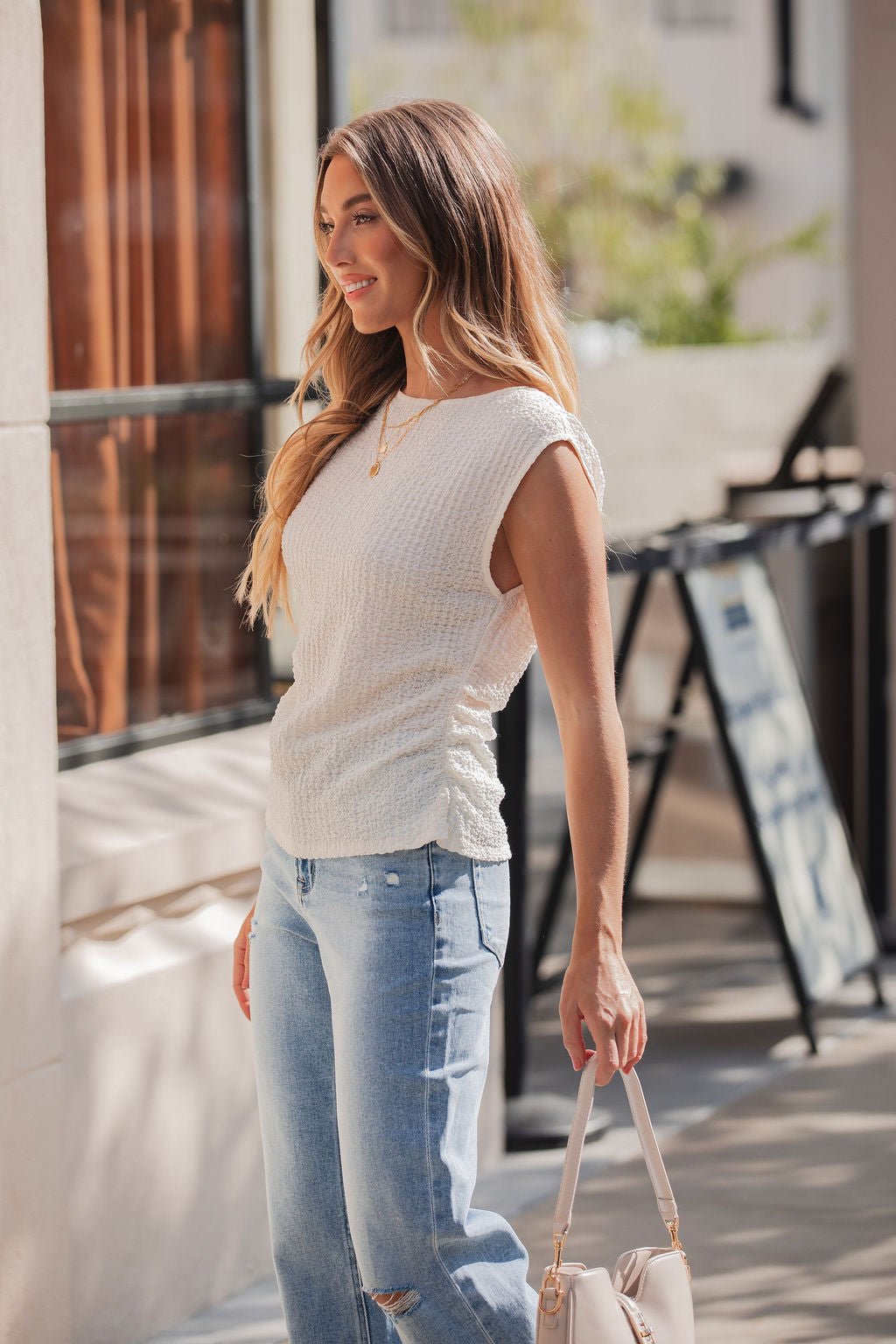 Woman with long hair in an Ivory Textured Cap Sleeve Top | Pre Order, light blue jeans, holding a handbag and smiling outdoors.