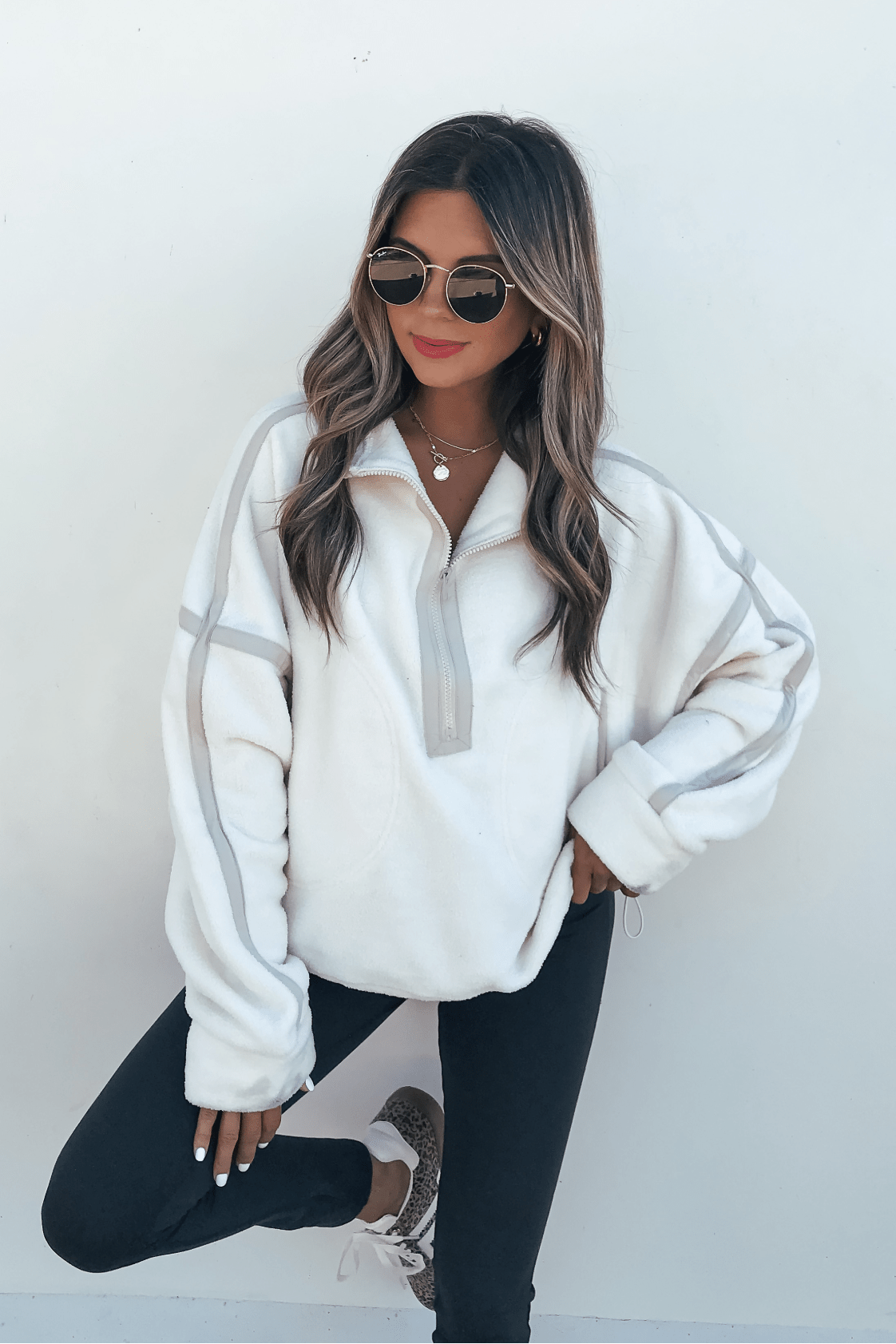 Woman in the Ivory Trim Detail Half Zip Sweatshirt, sunglasses, and dark pants poses by a white wall with red lipstick and wavy hair.