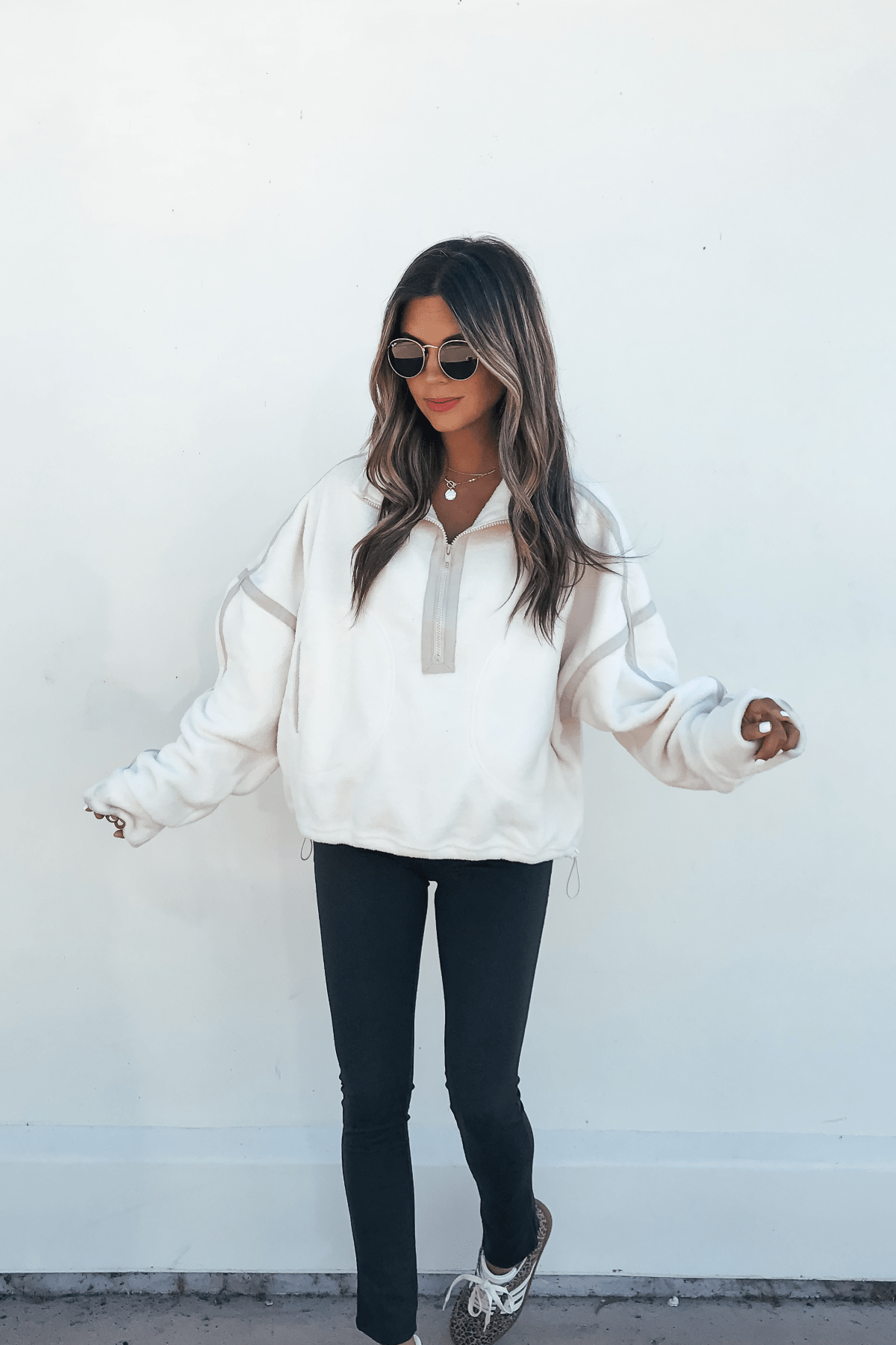 Woman in sunglasses and Ivory Trim Detail Half Zip Sweatshirt, black pants, and sneakers stands against a plain white wall.