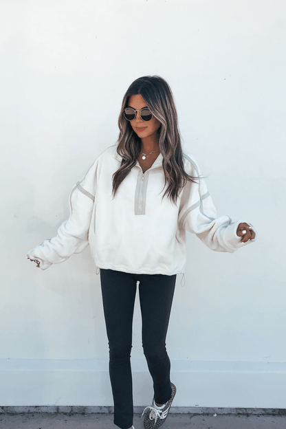 Woman in sunglasses and Ivory Trim Detail Half Zip Sweatshirt, black pants, and sneakers stands against a plain white wall.