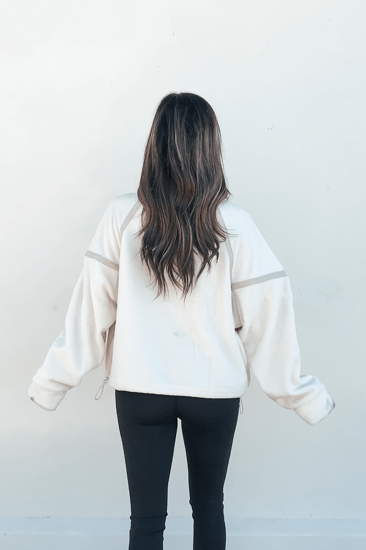 A woman with long, wavy brown hair wears an Ivory Trim Detail Half Zip Sweatshirt and black leggings, facing a white wall.