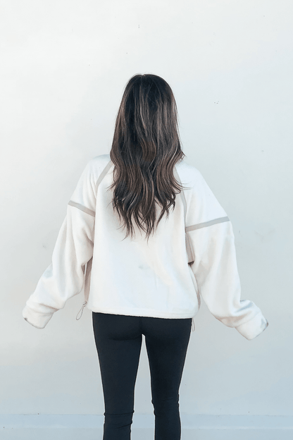 A woman with long, wavy brown hair wears an Ivory Trim Detail Half Zip Sweatshirt and black leggings, facing a white wall.