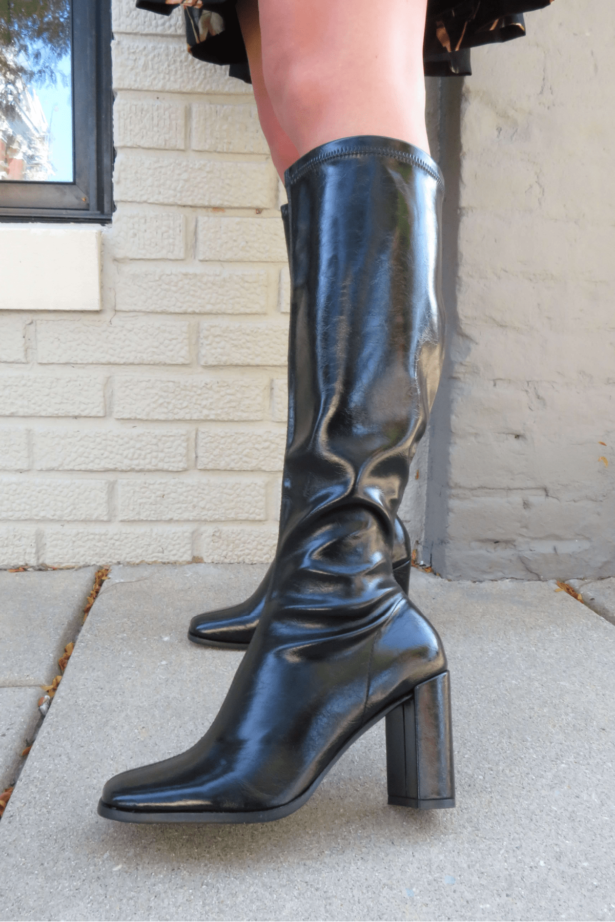 Someone wearing Ivy Black Faux Leather Heeled Knee High Boots stands on a concrete sidewalk by a brick wall.