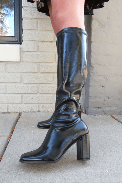 Someone wearing Ivy Black Faux Leather Heeled Knee High Boots stands on a concrete sidewalk by a brick wall.