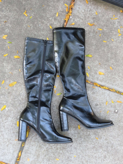 The Ivy Black Faux Leather Heeled Knee High Boots with zippers rest on concrete, surrounded by scattered yellow leaves.