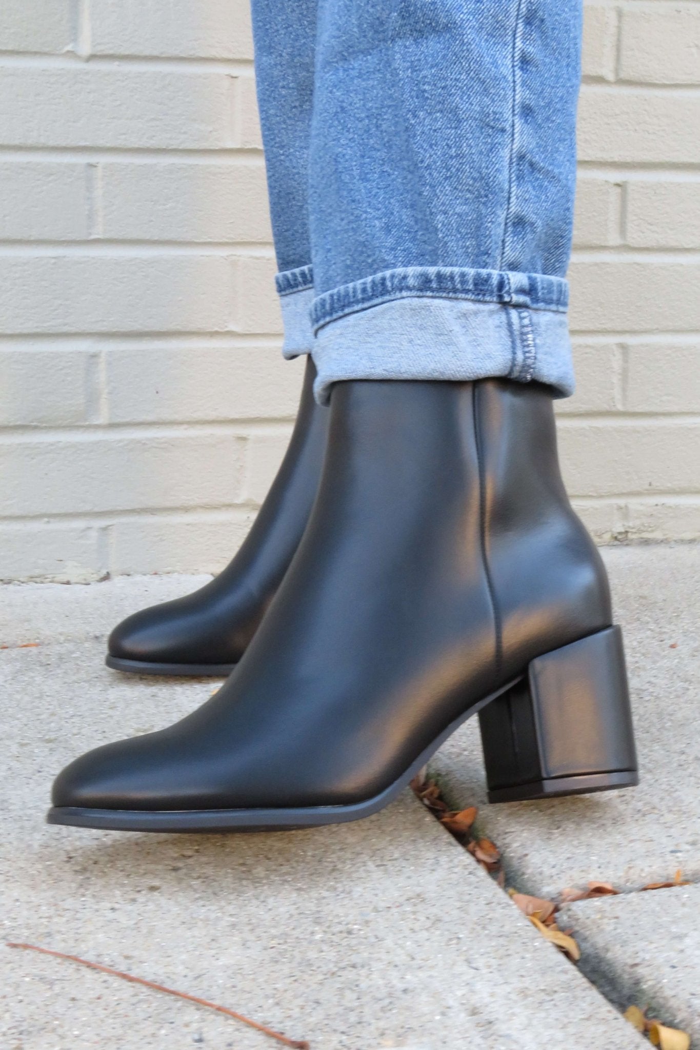 Person wears cuffed blue jeans and Jalen Black Faux Leather Booties, standing on a concrete sidewalk by a brick wall.