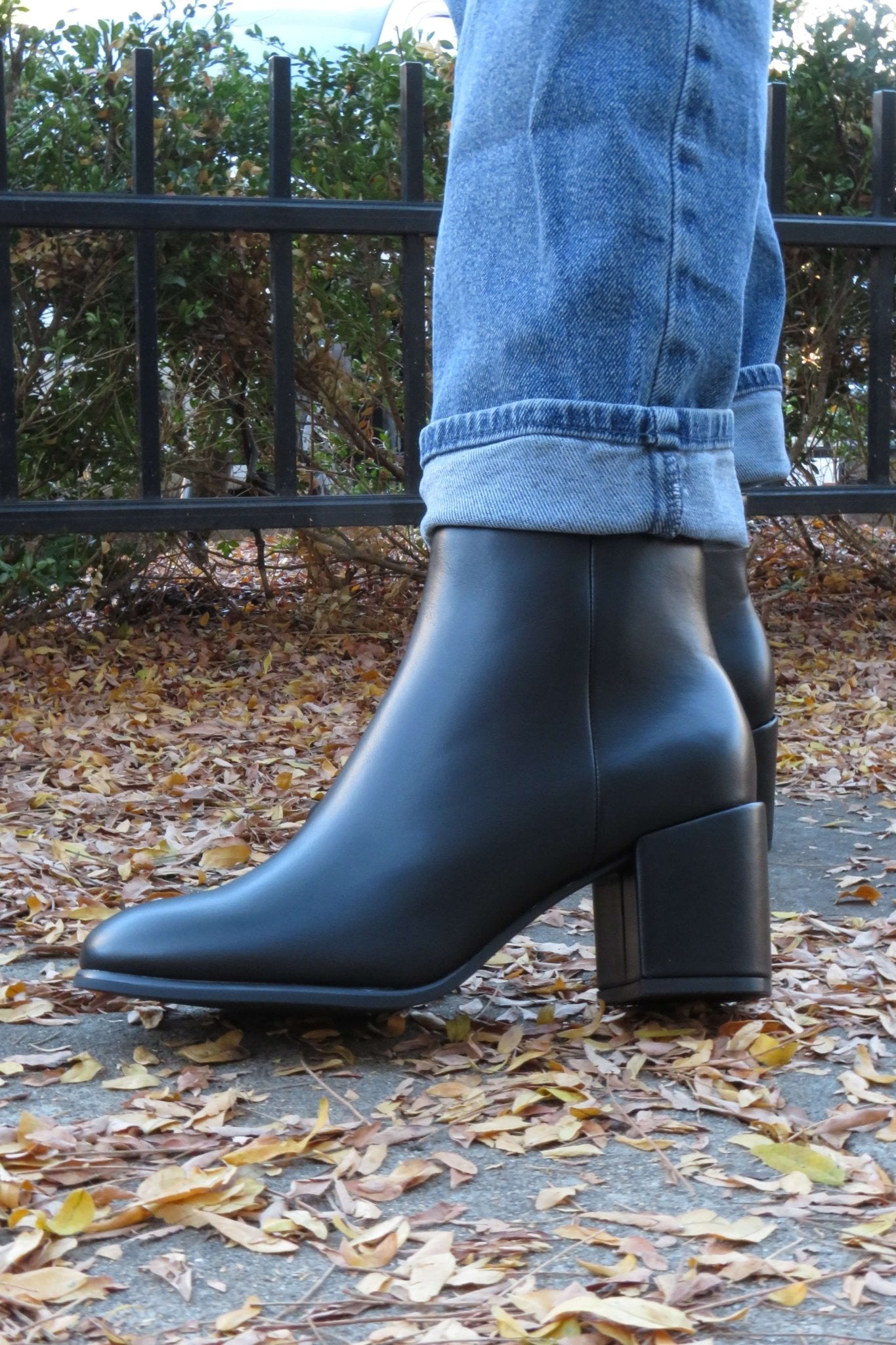 Someone in blue jeans and Jalen Black Faux Leather Booties stands on a leaf-covered sidewalk by a black metal fence.