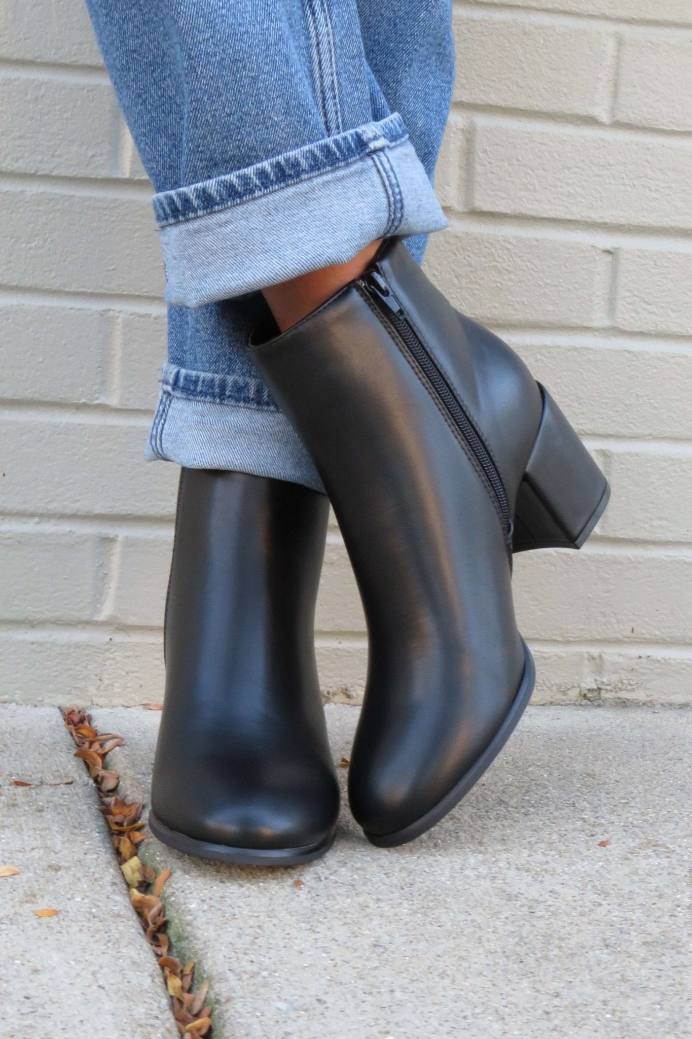 Person in cuffed blue jeans and Jalen Black Faux Leather Booties, standing on a sidewalk by a light brick wall.