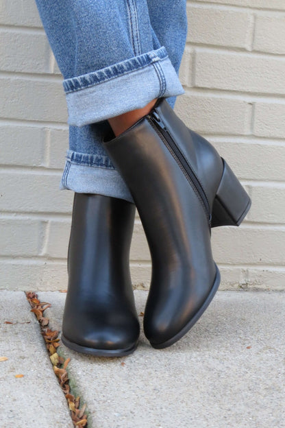 Person in cuffed blue jeans and Jalen Black Faux Leather Booties, standing on a sidewalk by a light brick wall.
