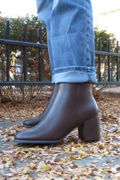 A person in cuffed blue jeans and Jalen Brown Faux Leather Booties stands on a leaf-covered sidewalk near a black iron fence.