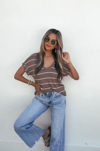 Woman in sunglasses wears the Jessica Striped Top - Mocha and wide-leg jeans, posing stylishly against a white wall.
