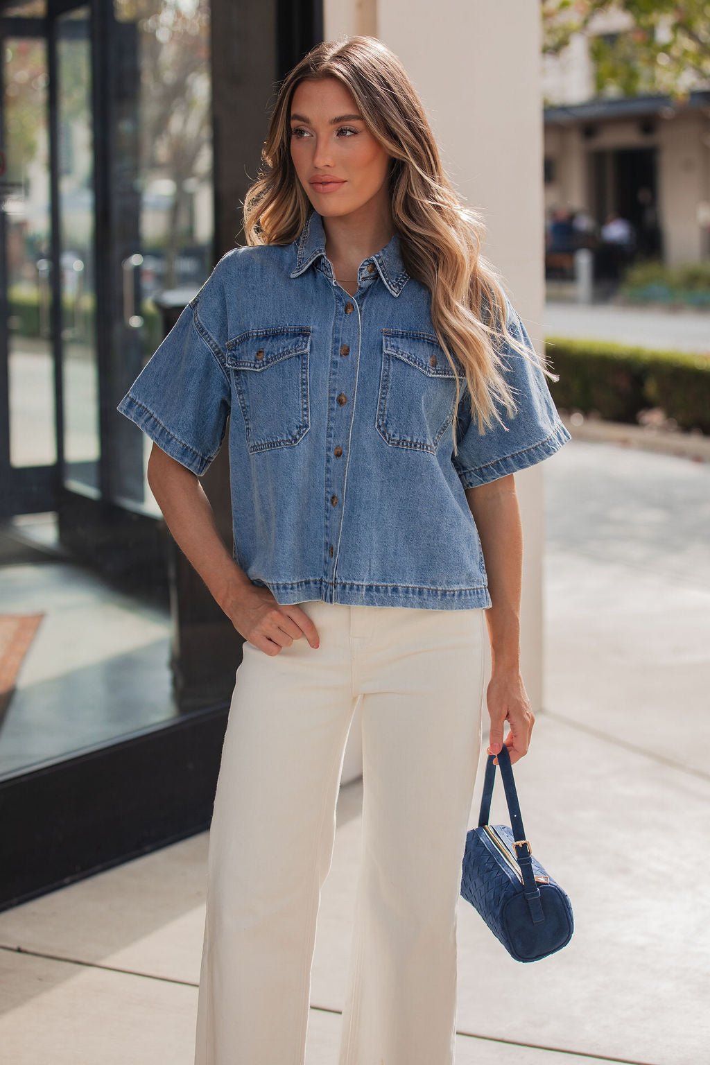 Wearing the Jessie Denim Button Up Shirt and white pants, a woman stands outside holding a small blue handbag.