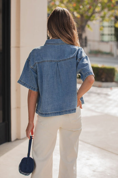 A woman with long hair wears the Jessie Denim Button Up Shirt and white pants, holding a small blue bag outdoors.