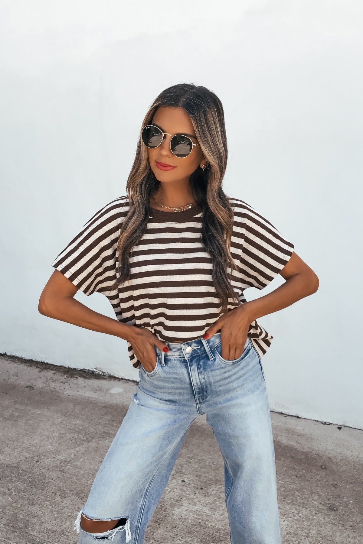 Woman in sunglasses and the Joan Striped Knit Top - Brown with ripped jeans stands confidently against a plain white wall.
