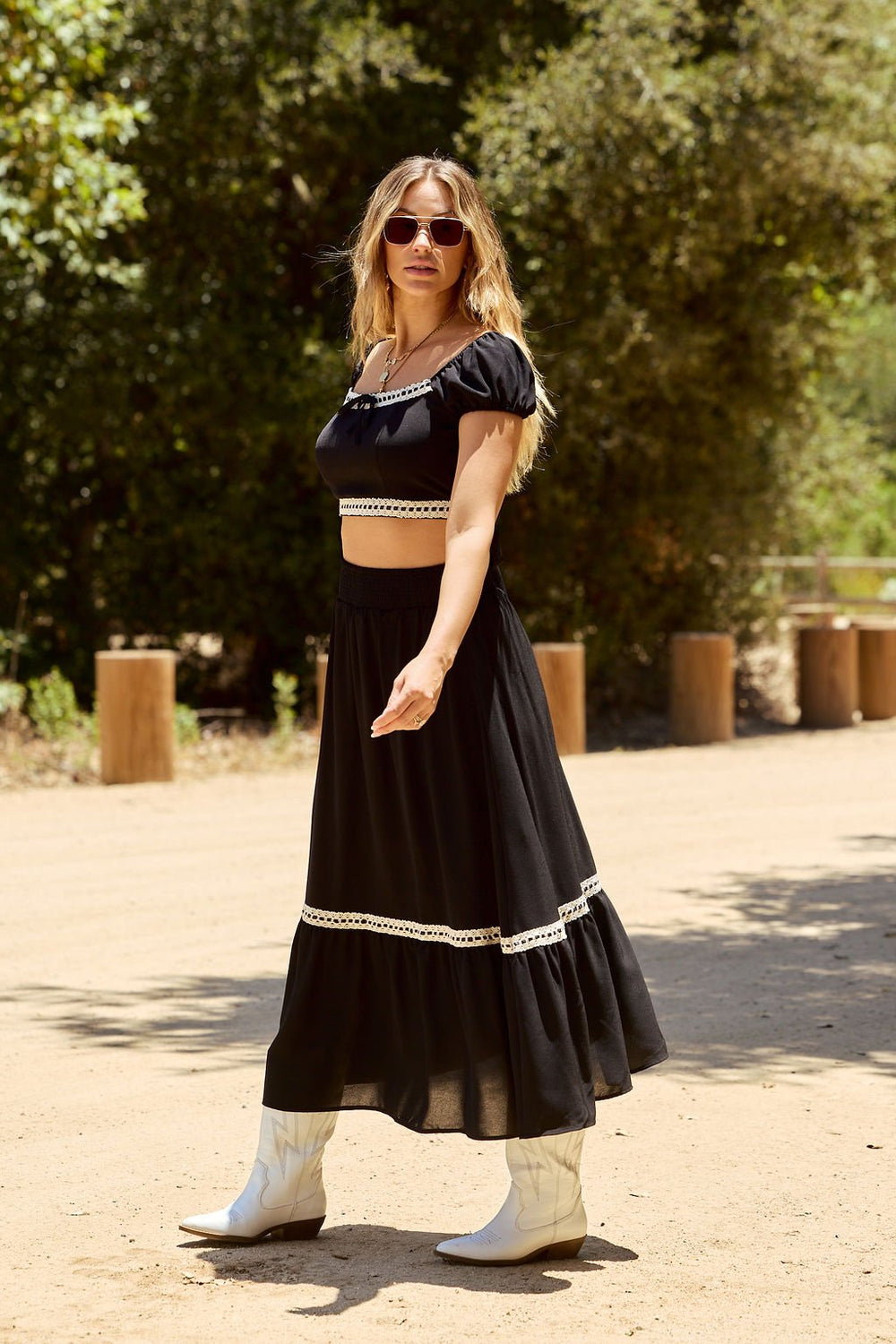Woman in the Jolene Black Lace Trim Maxi Skirt, white cowboy boots, and sunglasses, walking outdoors on a sunny day.