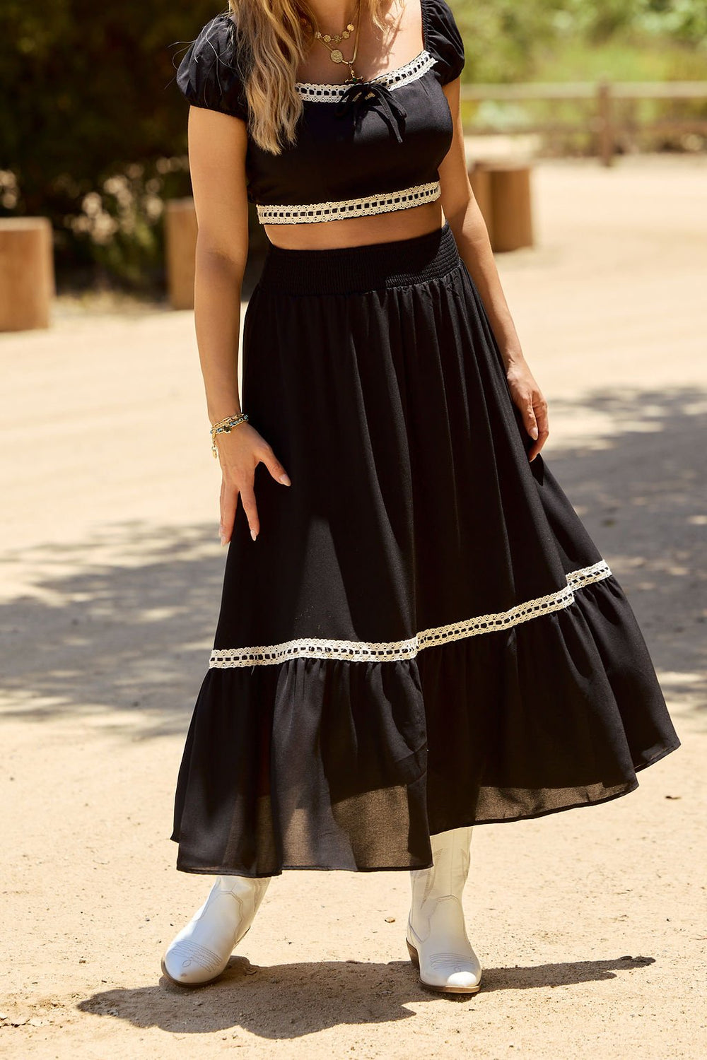 Woman in Jolene Black Lace Trim Maxi Skirt, black crop top, and white ankle boots, standing outdoors.